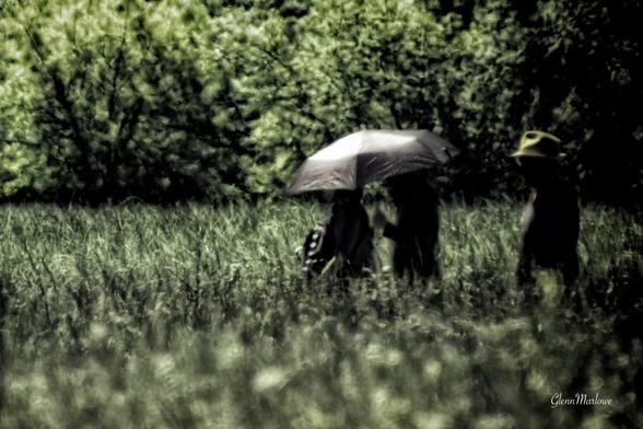 A blurred outdoor scene featuring two figures under a black umbrella in a grassy field, surrounded by lush greenery. A third figure stands nearby, wearing a hat.