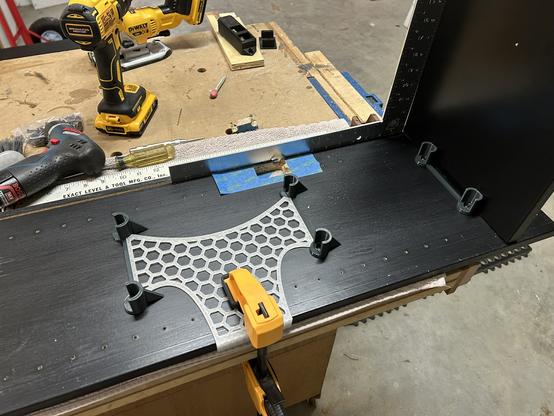 One side of an IKEA BILLY bookcase is clamped to a workbench.  The BILLY bottom is attached and clamped onto a carpenter's square.  Three FilaBilly mounting brackets are in place, and the FilaBilly alignment jig is between two of them.   One side is clamped to the BILLY's front edge.  The other two are nestled between two of the brackets.