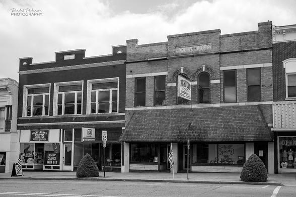 Black and white image of adjacent two story buildings facing the main street of a small town, one of them housing a hardware store.