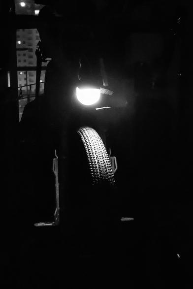 Photo noir et blanc, de nuit donc très sombre. Plan assez serré de l'avant d'un vélo dont le phare ressort très blanc et éclaire le pneu avant, large et sculpté. Le vélo est en hauteur sur un pied d'atelier. La lumière du phare est la seule source de lumière. À l'arriere plan un faible éclairage arrive d'une fenêtre donnant sur des immeubles de nuit