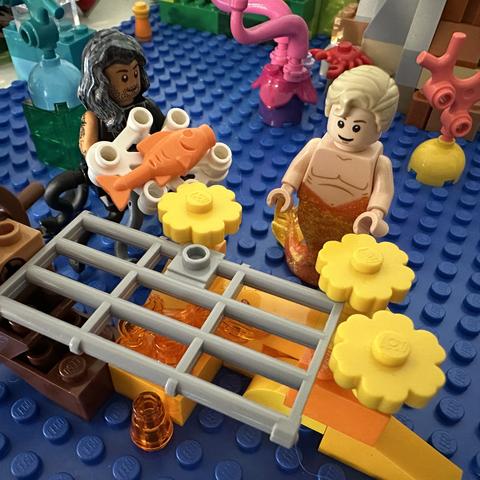 Lego MerKraken Ed offering an orange fish to Lego MerStede. In front of them, a cell door rests on some coral.