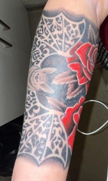 At the top of my forearm, from wrist to elbow.
PD: Tattoo on a forearm featuring a central bat with outstretched wings. The bat is rendered in shades of black and gray, with its wings forming a lacy, web-like pattern filled with intricate, decorative details. Surrounding the bat are two large, vivid red roses, one above and one below the bat, adding a bold splash of color to the otherwise monochromatic design. The tattoo covers a significant portion of the forearm and has a gothic, ornate style. The background includes some shading to give depth and contrast to the main elements.