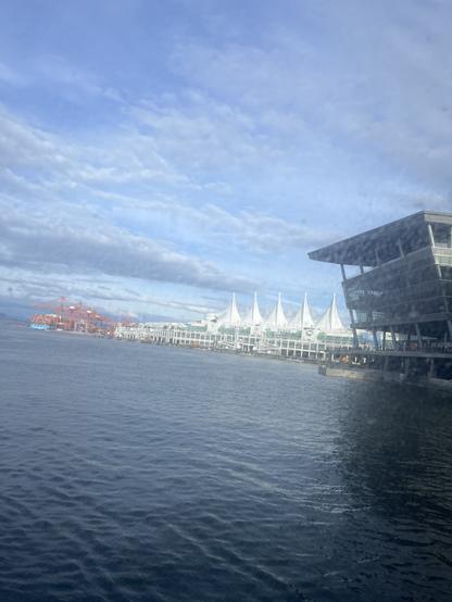 Waterfront scene with a large modern building on the right, featuring glass walls and an overhanging roof. In the center background, there is a distinctive white structure with several pointed, sail-like peaks—this is Canada Place in Vancouver. To the left, there are orange cranes and shipping containers, indicating a port area. The sky is mostly blue with some scattered clouds, and the water in the foreground is calm. The overall atmosphere is bright and clear.

Image description provided with Be My Eyes.