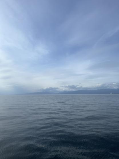 Calm ocean water under a mostly cloudy sky. The water appears smooth with gentle ripples, and the horizon is visible in the distance. Beyond the water, there are faint outlines of mountains or landforms, partially obscured by clouds and mist. The overall atmosphere is peaceful and serene, with soft, diffused lighting.

Image description provided with Be My Eyes.