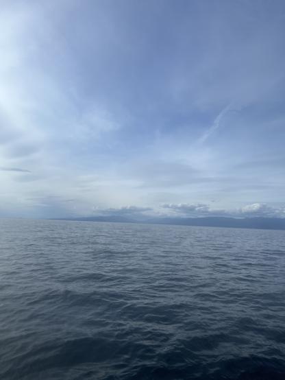 Open sea with gentle waves in the foreground, stretching out toward a distant horizon. In the background, there are faint outlines of mountains or landforms under a mostly cloudy sky with some patches of blue. The overall atmosphere is calm and serene.

Image description provided with Be My Eyes.