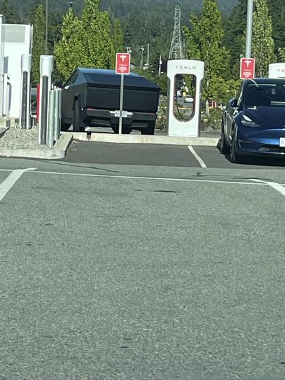 a picture across a parking lot toward a bank of tesla chargers is focused on a matt black cybertruck backing into a spot. The back is flat