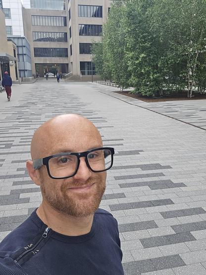 A selfie of me on a patterned grey stone path in front of a brutalist building, with birch trees on the right. I'm a bald, middle-aged, white man with a red beard flecked with white. I'm wearing glasses with thick black rims and a black shirt with a zipper that runs along the top right to the collar.