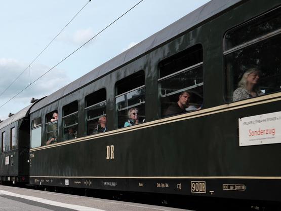 Ein alter Eisenbahnwagen, vom Bahnsteig aufgenommen aus niedriger Position und in schrägem Winkel. In den Fenstern sind Menschen zu sehen: vertieft in Gespräch, nach unten oder zum Fotografen blickend und in einem Fall auch schlafend. An der Seite prangt das Kürzel der Deutschen Reichsbahn „DR“ und ein Schild mit der Aufschrift „BERLINER EISENBAHNFREUNDE – Sonderzug“.

An old railroad car, taken from the platform from a low position and at an angle. People can be seen in the windows: engrossed in conversation, looking down or towards the photographer and, in one case, sleeping. The German Reichsbahn abbreviation “DR” and a sign with the inscription “BERLINER EISENBAHNFREUNDE - Sonderzug” are displayed on the side.