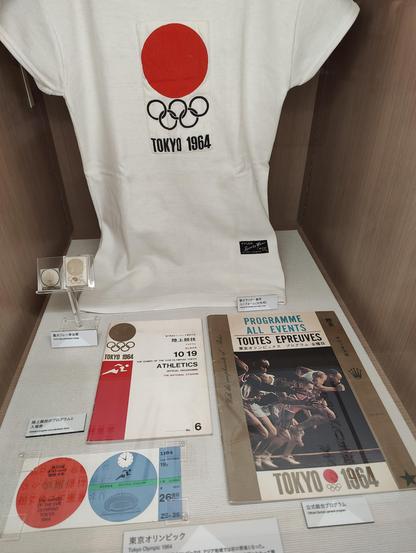A photo of a collection of memorabilia from the Tokyo 1964 Olympics in a museum exhibit, including a program, shirt, and other items