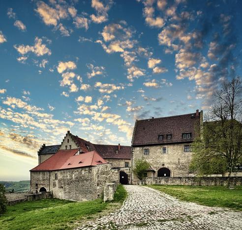A scenic view of a historic castle complex with stone walls and red-roofed buildings. A cobblestone path leads toward the entrance, surrounded by greenery and an expansive sky filled with clouds at sunset.