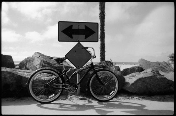 black abd white photography of a bike parked on a signe with a double ended arrow. there are rock behind the sign and then the beach. a palm tree trunk is visible also.