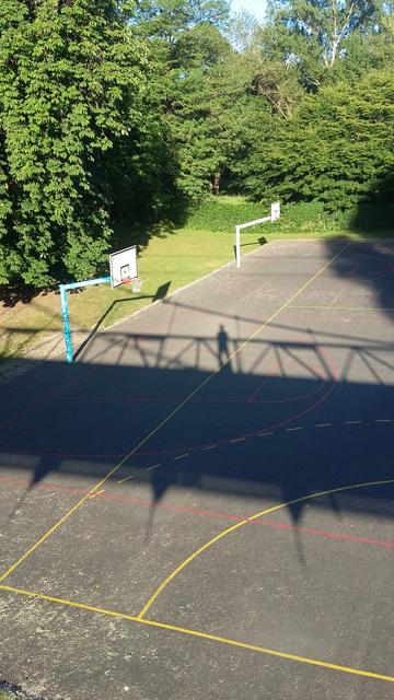 Vus d'en haut deux terrains de basket sur lesquels se projette l'ombre d'une passerelle. Sur celle-ci on distingue l'ombre de ma silhouette prenant cette médiocre photo avec mon sac à pain-du-vendredi à mes pieds.