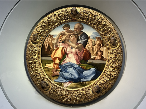 A woman in blue robes, muscular arms, and seemingly short hair, holds a baby on her right shoulder, and is embraced from behind by an older man with a white beard. Other secondary characters nearby, including presumably a young Saint Paul holding a cross ex tempore – the bay is Jesus and wouldn’t be crucified for another 30 years. The painting canvas is circular, with its frame being richly decorated in golden and including 5 small heads of key figures in Christian liturgical art.