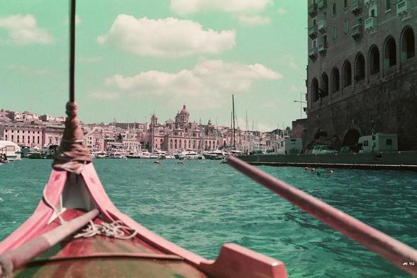 Farbfoto, aufgenommen auf einer Bootsfahrt im Hafen von Valletta, Malta. Eine gewisse Ähnlichkeit mit Venedig ist vorhanden: Gondel, Wasser stattliche Bauten am anderen Ufer. Unter blauem Himmel mit ein paar weißen Wolken. Farbstich grün-rosa durch den verwendeten Purple-Film. Leica R5 with 50 mm Summilux.