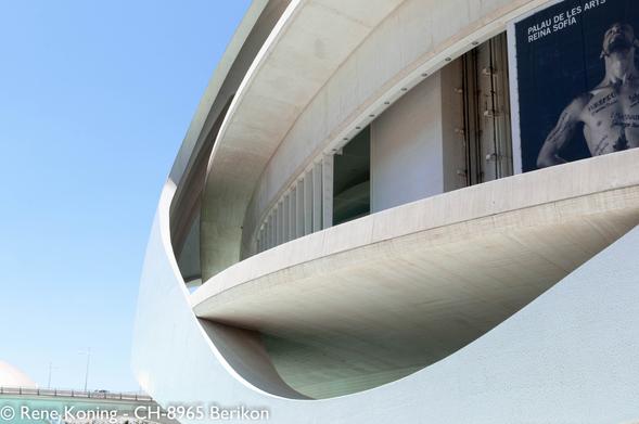 The image features a modern architectural structure, likely a museum or cultural center, with a distinctive curved design. The building's facade is predominantly white, with smooth, flowing lines that create a sense of movement and fluidity. The structure is composed of concrete, with a large, sweeping curve that forms a prominent part of the building's exterior. The upper section of the building has a large, dark poster with the text "PALAU DE LES ARTS REINA SOFIA" and an image of a shirtless man with tattoos. The sky is clear and blue, indicating a sunny day. The building's design is contemporary, with a minimalist aesthetic that emphasizes form and function. The image captures the building from a low angle, highlighting its impressive scale and architectural features. It is situated in Spain, city of Valencia