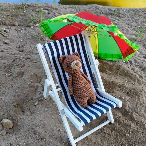 A picture of a small crocheted bear in a tiny doll sun chair with a parasol beside it, on some sand.