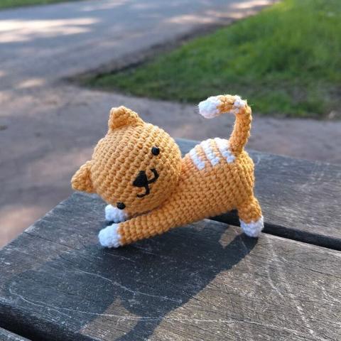 A picture of a crocheted amigurumi cat. It's orange-white colored, standing in playful pose, and looking sideways towards the viewer.