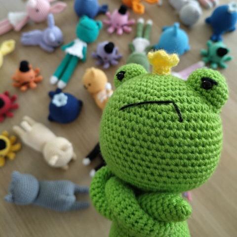 A picture of a crocheted frog wearing a crown, looking grumpy towards the viewer. In the background is a table full of colorful amigurumis.