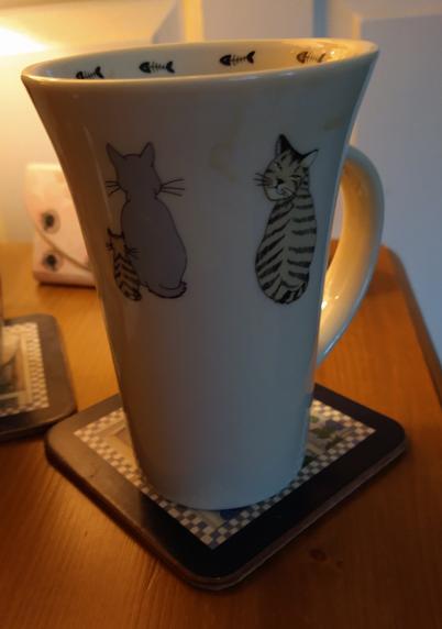 Mug of tea on a bedside cabinet with pictures of two grey cats one with it's back to us, and one looking over it's left shoulder