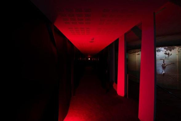 Image: Kate McElroy: installation shot; photo Roland Paschhoff | we are looking down an infra-red-lit dark corridor; on the left there appears to be a black wall or curtain, while to the right there are gaps  in the wall / white curtains through which we se a space in darkness except for a projection onto a wall or screen; the image is hard to decipher full, but it seems to be of a landscape of water and broken remnants of tree trunks and patches of land • “Kate McElroy is a Cork-based artist whose practice spans across media, presenting a dynamic constellation of work including expanded photography, moving image, sound, spoken word and sculptural installation. The work questions the altering environment and our place within it. Kate graduated with a First Class Honours Master’s Degree in Art and Process, MTU Crawford, 2021. Since graduating she has exhibited in over twenty national and international exhibitions and co-founded the artist collective inter_site. Recent exhibitions and…”