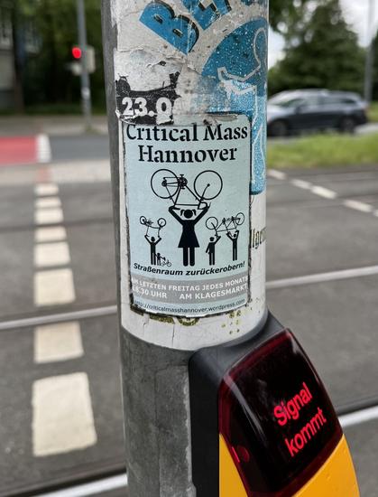 Foto. Ein hellblauer Aufkleber auf einem Ampelmast. Mit schwarzer Schrift und mehreren schwarzen gezeichneten Menschen, die Fahrräder in die Höhe halten. 
Text: 
Critical Mass Hannover
Straßenraum zurückerobern!
Am letzten Freitag jedes Monats
18:30 UHR
am Klagesmarkt,
http://criticalmasshanover.wordpress.com
