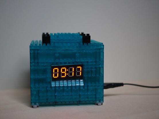 Eine Digitalanzeige in einem würfel-förmigen Gehäuse aus türkisen Klemmbausteinen zeigt die Uhrzeit an (09:17).