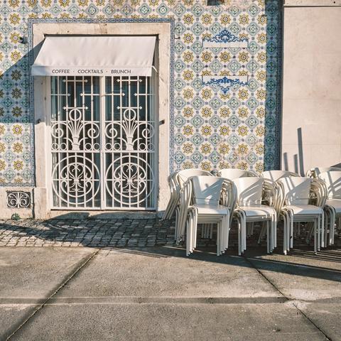 A cafe storefront with ornate white iron grillework over glass doors displaying "COFFEE • COCKTAILS • BRUNCH" signage. The building is covered in decorative blue and yellow Portuguese-style azulejo tiles in a repeating geometric pattern. Stacks of white plastic chairs are positioned to the right of the entrance. The ground features cobblestone paving with concrete sidewalk sections creating geometric lines.