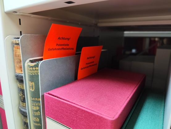 Foto: Bücher im Magazin, 2 davon mit rotem Zettel: "Achtung! Potentielle Gefahrstofffbelastung". DNB Leipzig.