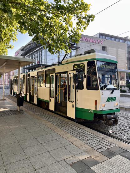Eine grün-weiße Straßenbahn hält an einer Haltestelle, deren Türen für Fahrgäste geöffnet sind. Es handelt sich um eine Tatra des Typs KT4D. In der Nähe ist im Hintergrund ein großes modernes Gebäude mit der Aufschrift "Cinemaxx" zu sehen, zusammen mit schattenspendenden Bäumen.