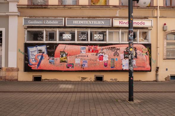 Foto von einem Laden für Gardinen, Heimtextilien und Kindermode. Das große Schaufenster ist hinter Spanplatten verschwunden. Auf den Spanplatten ein großes Graffiti mit einem Arm und einer Hand, die in eine Todo-Liste u.a. "AfD stoppen" schreibt. Diverse Zettel und Tags sind auf dem Graffiti. Das ganze Gebäude wirkt heruntergekommen.