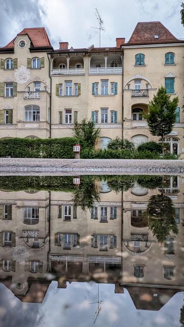 Das Bild zeigt ein dreistöckiges Gebäude mit einer detaillierten Architektur, das sich in einer Pfütze spiegelt. Das Gebäude hat mehrere Fenster mit grünen Fensterläden und einige Balkone. Die Fassade ist mit dekorativen Elementen verziert, darunter ein Relief einer biblischen Szene auf der linken Seite. Das Dach ist rot und hat mehrere Schrägen, mit einer großen Antenne darauf. Vor dem Gebäude befindet sich eine Hecke, und einige Bäume rahmen die Szene ein. Die Spiegelung im Wasser ist klar und fast perfekt, wodurch das Gebäude und seine Umgebung doppelt sichtbar erscheinen.