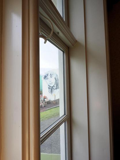 A view through a window showing a mural of a girl’s face on the exterior wall of a building. The scene includes grassy ground and playground equipment visible outside. The window frame is wooden, with a rope handle.