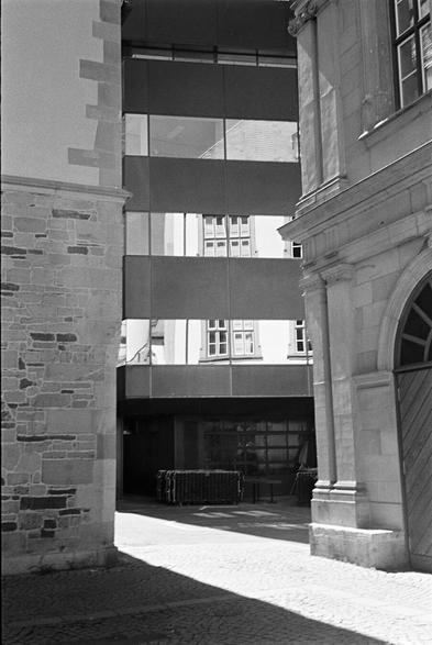 The uploaded image shows a narrow, sunlit pathway between two old, stone buildings, leading towards a modern, multi-story structure with large windows and horizontal panels. The building on the left is made of rough, irregular stone blocks, with a slightly textured, light-colored surface visible at the top. The building on the right, also stone, appears more ornate, with a rounded archway and a dark wooden door visible on its right side. The pathway itself is paved with cobblestones on the right and a darker, possibly asphalt or packed earth, surface on the left, where a long shadow stretches across it. In the background, the modern building dominates the view, its glass facade reflecting parts of the older buildings and the sky. Below the modern building, a dark, recessed area suggests an entrance or an underground level, with what appears to be stacked crates or furniture in front of it.
