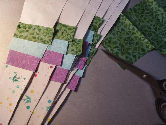 Photo of an ironing mat with fabric and scissor laying on top. On the left side lay several paper wedges with fabric in a rainbow order attached to them. On the right side lays green batik fabric with a big fabric scissor.
