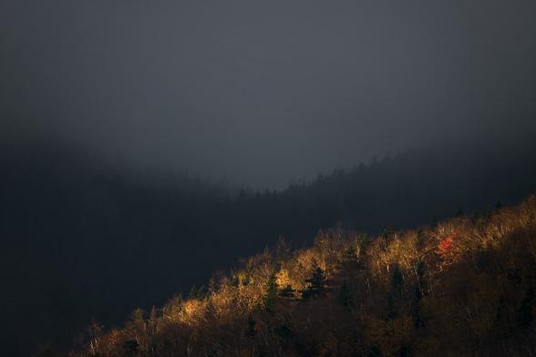 A mountain ridge catches a ray of golden sunshine in an otherwise dark monochromatic foggy landscape