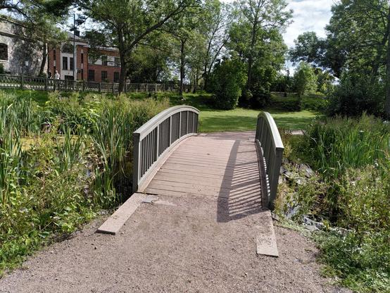 Eine Brücke in einer parkähnlichen Umgebung mit Wiese und Bäumen. In etwas Entfernung sieht man eine Häuserfront auf der linken Seite.
