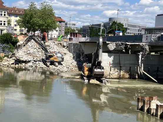 Das Bild zeigt die Abrissarbeiten an der Gänstorbrücke in Ulm. Im Vordergrund fließt die Donau, rechts stehen noch Reste der Brücke, 3 Bagger arbeiten sich am Schutt ab, im Hintergrund weitere Gebäude.