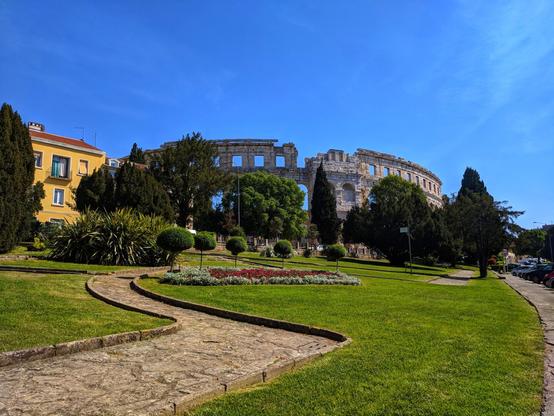 A wide, sunlit view of the Pula Arena in Croatia, framed by green parkland and a clear blue sky. The ancient Roman amphitheatre's weathered stone facade stands prominently in the mid-ground, partially obscured by mature trees. A winding stone pathway leads through a well-maintained lawn dotted with trimmed bushes and flowerbeds towards the arena. To the left, a colourful building with yellow and white walls and a red roof is visible amongst the trees. Parked cars line the right edge of the frame.