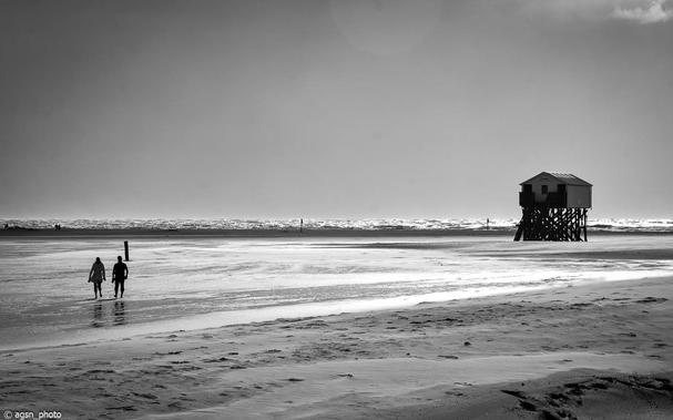 Ein Schwarz-Weiß-Bild. Ein breiter Sandstrand bei Ebbe, links im Vordergrund ein Paar, das aus dem Bild läuft, weiter rechts in der Ferne eine große Holzhütte auf Stelzen. /
A black-and-white image. A wide sandy beach at low tide, a couple walking out of the frame in the left foreground, and further to the right in the distance, a large wooden hut on stilts.