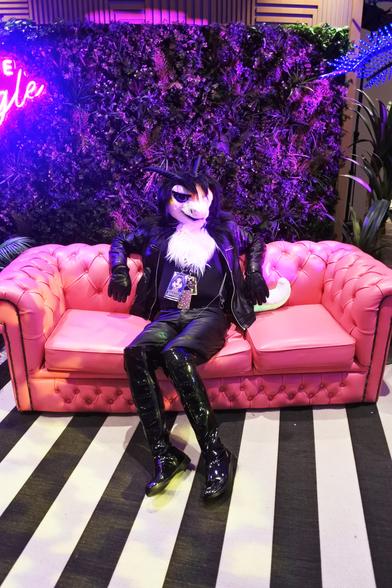 Fursuit photo - a furred dragon wearing leather jeans, jacket and gloves sits on a faux-leather couch in one of the photoshoot areas.