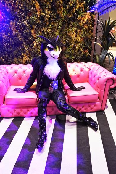 Fursuit photo - a furred dragon wearing leather jeans, jacket and gloves attempts a sexy pose on a faux-leather couch in one of the photoshoot areas.