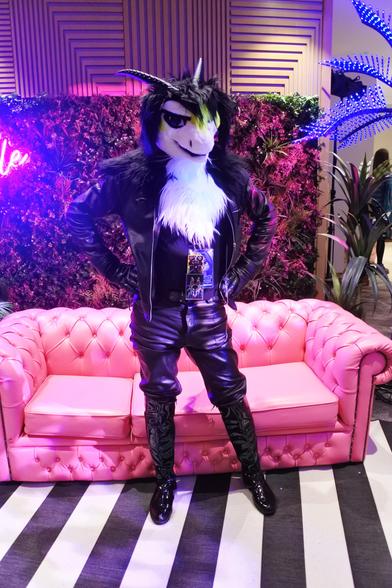 Fursuit photo - a furred dragon wearing leather jeans, jacket and gloves stands in front of a faux-leather couch in one of the photoshoot areas.