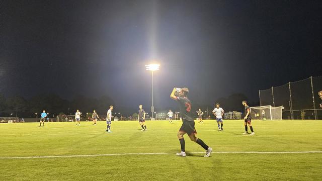A player in a black kit with red numbers takes a throw-in during a soccer match under the lights.