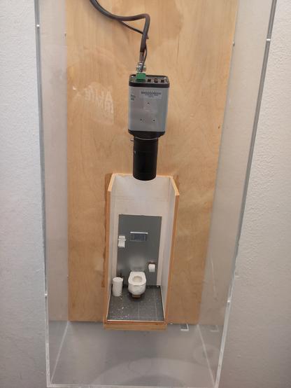 In einem klaren Plexisglas-Kasten wird das maßstabs-getreue Modell einer Klo-Kabine von oben abgefilmt.