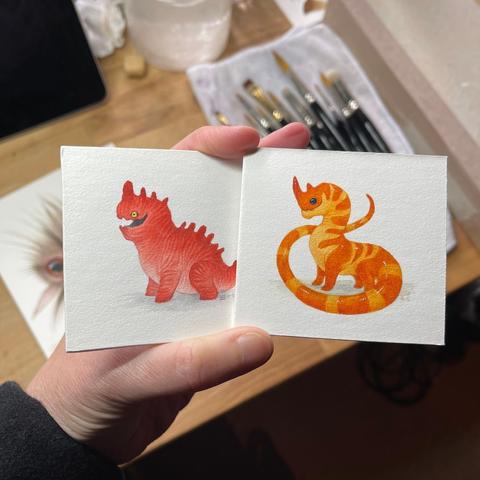 a photo of my hand holding two tiny dragon paintings, one is red and stripy and the other is bright orange and rich yellow. in the background you can see my paintbrushes laid out and a hint of a painting i've been working on which appears to be a creature with long whiskers and a large eye.