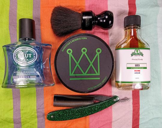 (Le Chat Mistral description)

The image showcases a collection of grooming items arranged on a striped background. At the top left, there's a bottle of Brut Oceans aftershave. To the right of the aftershave, there's a black shaving brush with a wooden handle. Below the brush, there's a round container of Barrister's Reserve Fern luxury shaving soap. To the right of the shaving soap, there's a bottle of Stirling Soap Co. Varen aftershave splash. At the bottom of the image, there's a green straight razor with a decorative handle. The items are neatly laid out, creating a visually appealing arrangement.