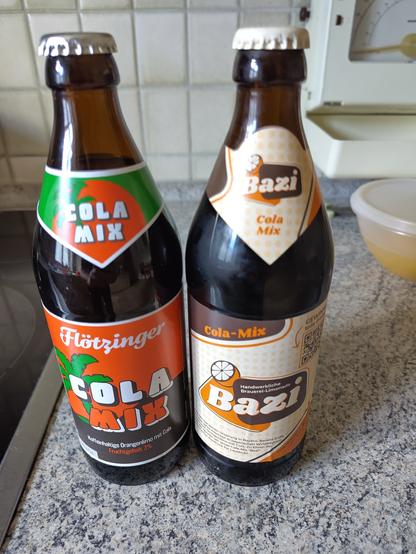 2 Flaschen Spezi auf einer Küchenarbeitsplatten Unterlage.
Lima Flötzinger Cola Mix, das Label in orange mit Aufdruck 3%Frucht und rechts das Bazi Spezi. im gelb mit leichten orange Tönen.