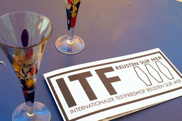 Flyer mit Aufschrift "ITF – Internationaler Testfriedhof Reusten Sur Mer" und drei symbolisierten Särgen.

Blaue Tischplatte mit eben diesem Flyer drauf und zwei bunten Miniatur-Sektkelchen.