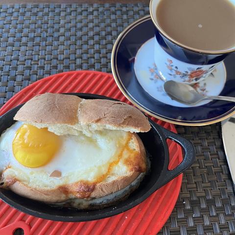 A yellow egg yolk and cooked white over a slice of cheese on hallowed roll served in a small oval cast iron skillet, accompanied by a cup of coffee in a decorated cup and saucer.