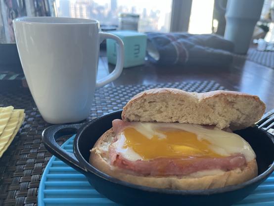 A white coffee cup, a small cast iron skillet containing a dinner roll sandwich with ham, cheese, and a sunny-side-up egg, on a blue silicone mat. In the background, there is timer cube, pot holder and a glimpse of a cityscape.